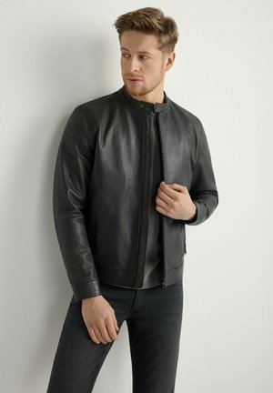 Czarna skórzana kurtka z kołnierzem typu stójka i zamkiem błyskawicznym, o gładkiej teksturze i dopasowanym fasonie. Noszona na ciemnej bluzce i dżinsach.