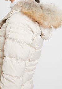 Giacca a vento beige con cappuccio di pelliccia rimovibile, con pannelli cuciti e logo Calvin Klein Jeans sulla manica sinistra. Texture liscia.