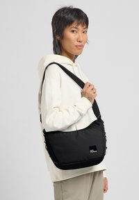 Borsetta nera a tracolla con superficie texturizzata, tracolla regolabile e patch con logo. Presenta un design semplice e materiale morbido e resistente.