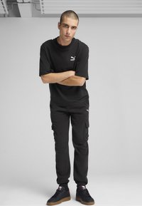 Schwarzes Puma-T-Shirt und Cargo-Hose aus leichtem Material. Enthält das weiße Puma-Logo. Schwarze Sneakers mit einer Gummisohle und roten Akzenten.