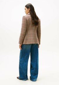 Tweedblazer met een gestructureerde pasvorm in beige, rood en zwart patroon, gecombineerd met losse blauwe spijkerbroek. Lang steil haar zichtbaar.