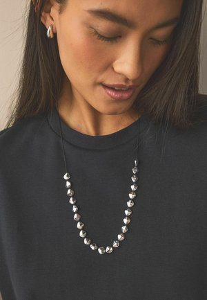 Frau mit langen dunklen Haaren und geschlossenen Augen, die silberne Creolen und eine silberne Perlenkette über einem schwarzen Oberteil trägt.