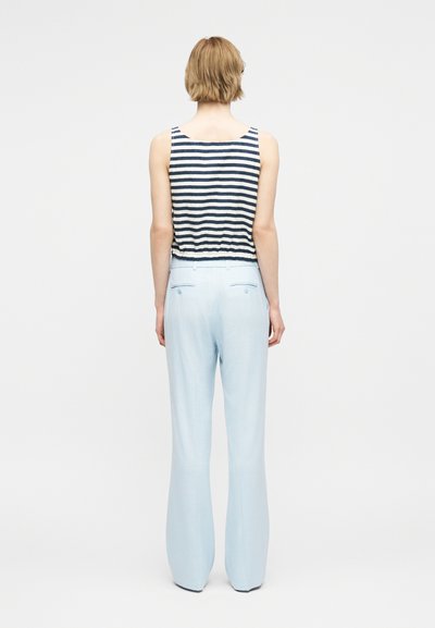 Γυναίκα που στέκεται με την πλάτη, φοράει αμάνικη μπλούζα με μπλε ναυτικό και λευκές ρίγες και ανοιχτό μπλε παντελόνι φαρδιάς γραμμής σε απλό φόντο.