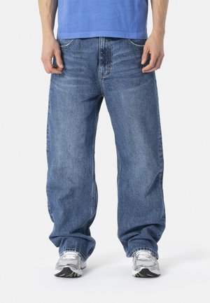 Homme portant un jean bleu ample, des baskets blanches et une chemise bleue rentrée, debout devant un fond clair uni.