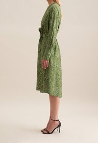 Zelené midi šaty s bílým puntíkovaným vzorem, dlouhými rukávy a páskem v oblasti pasu. Spárováno s černými botami na vysokém podpatku s řemínky. Hladký textilní povrch.