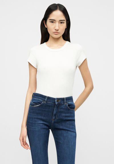 Νεαρή γυναίκα με ίσια σκούρα μαλλιά φοράει εφαρμοστό λευκό κοντομάνικο μπλουζάκι και σκούρο μπλε ψηλόμεσο τζιν, στέκεται μπροστά σε απλό φωτεινό φόντο.