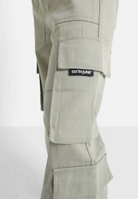 Pantalons cargo en vert clair, fabriqués en coton durable, avec plusieurs poches et une étiquette sur la cuisse gauche. Tissu texturé avec une sensation légèrement rugueuse.
