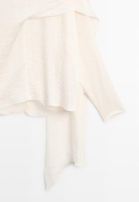 Lichtbeige, textuurvolle top met een asymmetrische zoom en lange mouwen, gemaakt van een zachte, vloeiende stof. Geen zichtbare merknamen of patronen.