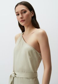 Jumpsuit leggero beige a spalla unica realizzato in tessuto morbido, con una tasca laterale e una cintura legata in vita per definire la silhouette.