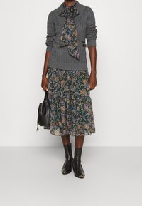 Žena ve šedém svetr s kabelovým úpletem přes květinové midi šaty, černé kotníkové boty s kovovými špičkami a držící černou kabelku.