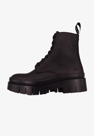 Botas de cuero negras hasta el tobillo con punta redonda, suela gruesa y texturizada, costuras prominentes y una lengüeta en la parte trasera para facilitar su puesta.