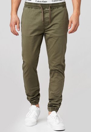 Man die olijfgroene joggers draagt met elastische boorden en een tailleband met trekkoord, gecombineerd met witte sneakers en een zichtbare tailleband van Calvin Klein ondergoed.