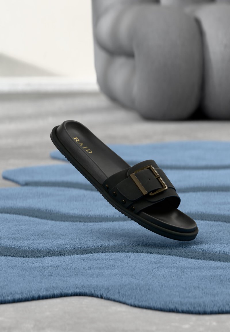 Sandale à glissière noire avec boucle dorée reposant sur un tapis bleu texturé, des coussins gris flous en arrière-plan.