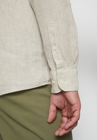 Beige Leinenhemd mit Knöpfen und Manschette, getragen zu olivgrünen Hosen. Die Textur des Stoffes ist sichtbar.