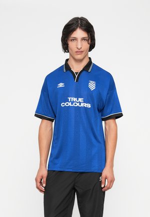 Joven con cabello oscuro que lleva una camiseta deportiva de color azul con cuello y pantalones negros, de pie contra un fondo blanco liso.