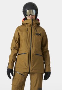 Traje de esquí de dos piezas marrón con capucha, que presenta cremalleras y bolsillos negros. Incluye guantes negros y un casco con visor espejado.