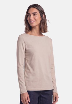 Femme aux cheveux mi-longs portant une chemise beige à manches longues et un pantalon foncé, souriante et regardant sur le côté.