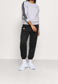 Persona vestida con una camiseta de manga larga gris con cinta de logo negra en las mangas, pantalones negros con logo, calcetines blancos y zapatillas blancas, de pie sobre un suelo gris.