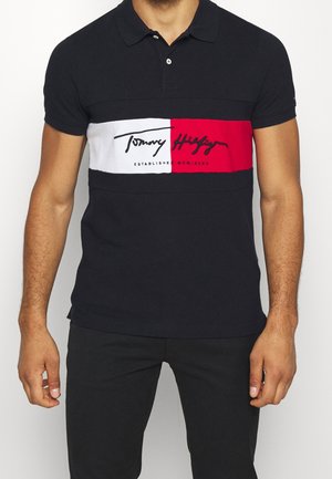 Man draagt een zwart poloshirt met witte en rode horizontale streep en de tekst "Tommy Hilfiger" op de borst, gecombineerd met een zwarte broek.