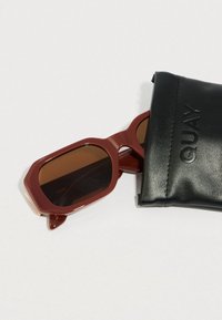 Rudos atspalvio, aštuonkampio formos akiniai nuo saulės su tamsiai raudonu rėmeliu, iš dalies gulintys juodos odinės dėklo viduje, kuriame įspaustas užrašas "QUAY".