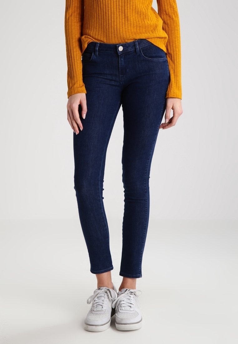 Femme portant un jean skinny bleu marine, un haut à manches longues côtelé moutarde, et des baskets blanches, debout devant un fond clair uni.