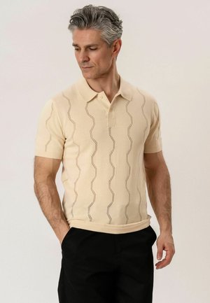Homme portant un polo à manches courtes beige avec des motifs en zigzag verticaux et un pantalon noir, debout les mains dans les poches, regardant vers le bas.
