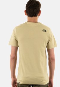 Světlobéžové tričko s krátkým rukávem vyrobené z bavlny, s malým černým logem na horní části zad. Hladký povrch, ležérní design.