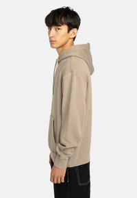Beige hoodie med framficka, dragsko på huvan och ribbade ärmslut. Buren med mörka jeans, vilket visar en avslappnad passform och mjuk textur.