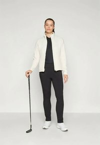 Svijetlosmeđa jakna na zip, crni polo majica, crne tajice i bijele cipele. Model drži golf štap, stojeći naslonjen na običnu pozadinu.