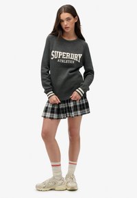 Un pull en tricot gris avec "SUPERDRY ATHLETICS" en blanc, associé à une jupe plissée à carreaux noir et blanc et des chaussures de sport beige avec des chaussettes rayées.