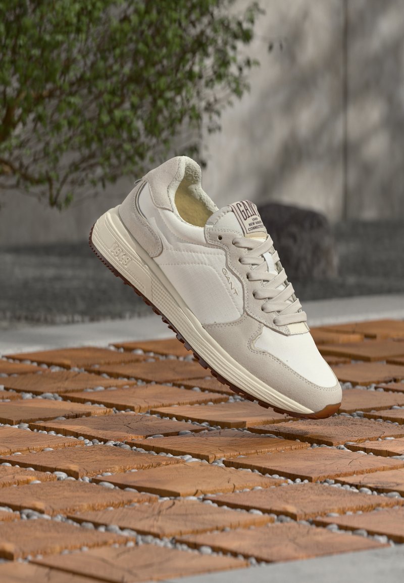 Μπεζ και λευκό αθλητικό παπούτσι με κορδόνια να αιωρείται πάνω από μια βεράντα με τετράγωνα ξύλινα πλακάκια με μικρά κενά γεμάτα χαλίκια, με πράσινο φυτό και πέτρινο φόντο.