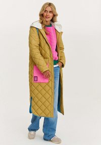 Dlhý prešívaný kabát v horčicovej žltej farbe s ružovou vreckom, bielou kapucňou podšitou fleeceom a bočnými rozparkami; nosený cez prúžkovaný ružový a zelený sveter.