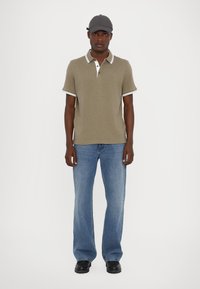 Olivengrønn polo med hvite detaljer, kombinert med lyseblå rette jeans og svarte sko. Modellen har på seg en grå caps.