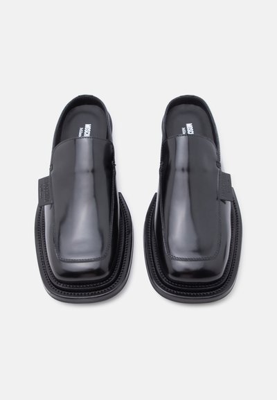 Chaussures en cuir noir brillant sans lacets, avec des bouts carrés et des semelles épaisses superposées, vues de dessus sur un fond clair uni.