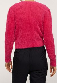 Chlupatý růžový svetr s žebrovanou texturou, dlouhými rukávy a zkráceným střihem, spárovaný s černými elastickými kalhotami. Žádné viditelné zapínání.