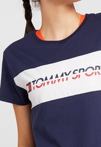 Navy blue cotton T-shirt with a white horizontal stripe featuring "TOMMYSPORT" in red and black. Short sleeves and crew neck design.