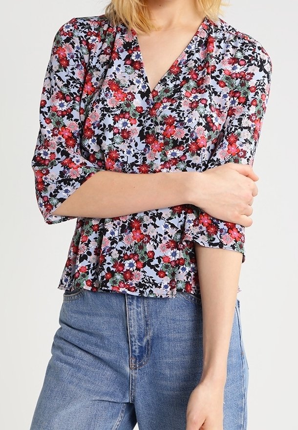 Blusa floreale in tessuto azzurro chiaro con motivo floreale rosso e verde. Presenta un collo a V, maniche a tre quarti e una vestibilità morbida.
