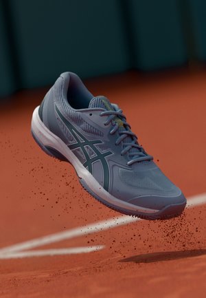 Shoe desportivo cinza claro com um superior de malha texturizada, acentuado por um logo cinza escuro e entressola branca, pairando acima de um court de argila.