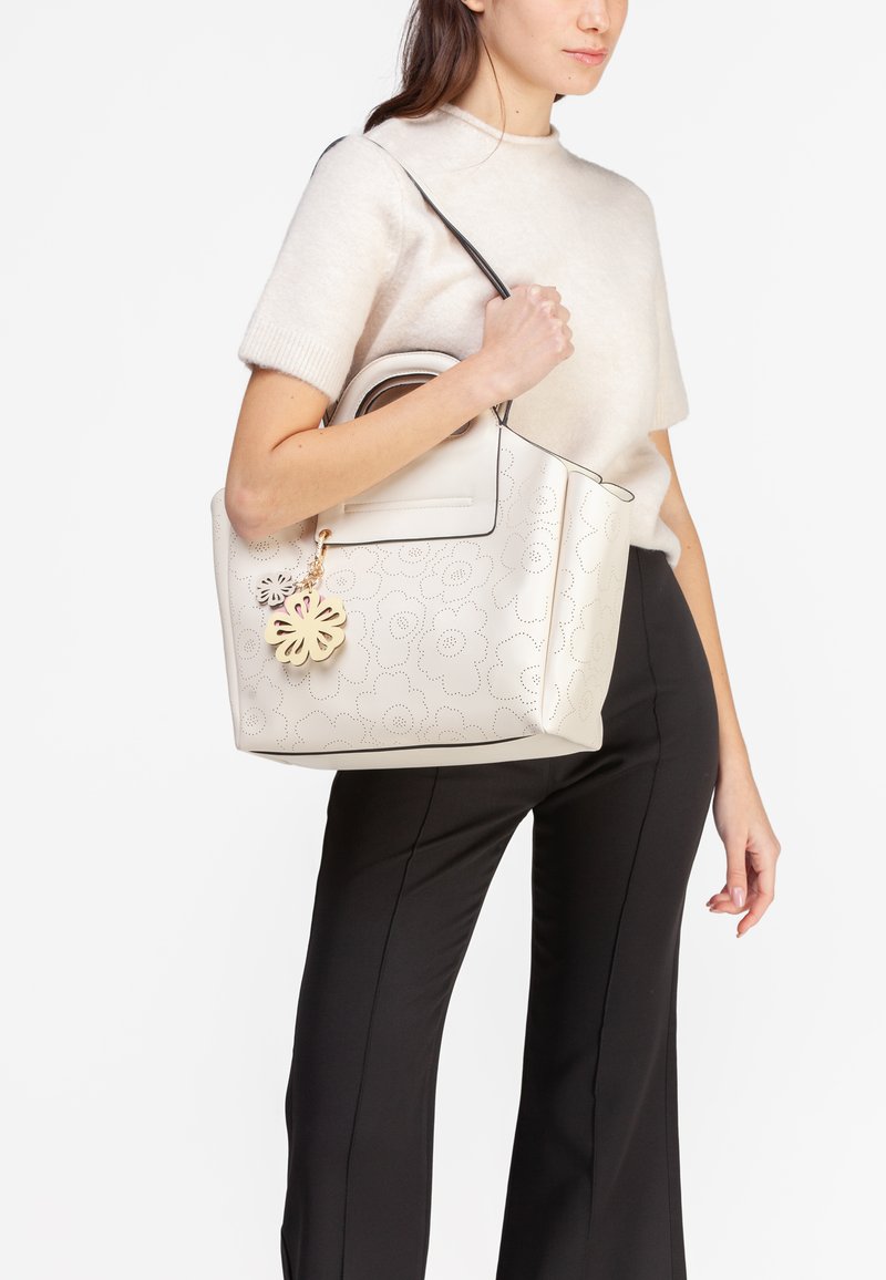 Beige håndtaske med blomstermønster i perforeret design, sort pynt og en dekorativ charm. Glat lædermateriale. Rummelig med dobbelte håndtag.