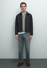 Sort letvægtsjakke, grå polo, hvid skjorte under, og falmede blå jeans. Brune snøresko. Enkel, moderne design.