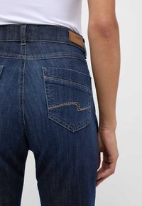 Mörkblå denimjeans med hög midja, som har en brun läderlapp på baksidan och två bakfickor med sömdetaljer.