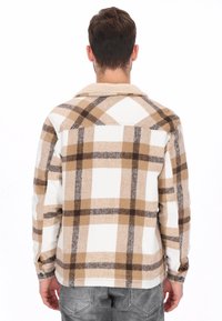 Camisa de franela en cuadros beige y blanco con cuello suave, mangas largas y puño abotonado. Tejido con textura y patrones de cuadros variados.