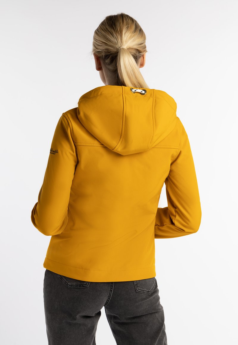 Chaqueta amarilla de softshell con capucha, con un detalle de logo en la parte trasera y textura suave. Combinada con unos pantalones gris oscuro, con hardware mínimo visible.
