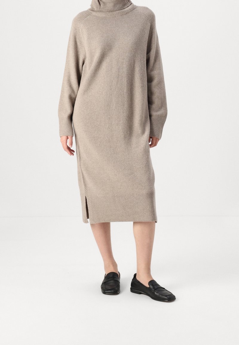 Personne portant une robe-pull beige à manches longues et col roulé avec une fente latérale, associée à des mocassins noirs slip-on, debout sur un sol blanc.