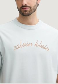 Jasnoniebieska koszulka z krótkim rękawem wykonana z miękkiej tkaniny, z beżowym, kursywnym napisem "calvin klein" wyhaftowanym na piersi.