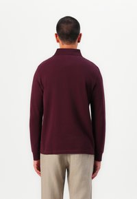 Burgundy long-sleeve polo shirt with a collar, fitted design, and ribbed cuffs, paired with light beige trousers. Simple texture, no patterns.