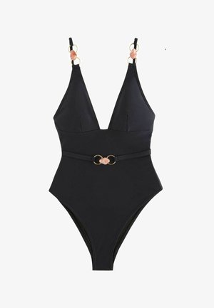 Femme aux cheveux mouillés se tient près de l'eau au coucher du soleil, portant un maillot de bain une pièce noir avec un décolleté profond et un détail de ceinture décorative.