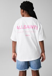 Balta per didelė t-shirt su rožiniu "ALLSAINTS" tekstu ant nugaros, derinama su šviesiai mėlynomis džinsinėmis šortais, turinčiomis nusivylusias kraštines.