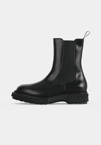 Botte Chelsea en cuir noir avec panneaux latéraux élastiques, semelle épaisse rainurée et languette à l'arrière sur un fond clair uni.