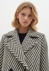Čierno-biely kabát v houndstooth vzore s dvojradovým zapínaním, veľkým klopovým golierom a čiernymi gombíkmi. Textúrovaná tkanina.